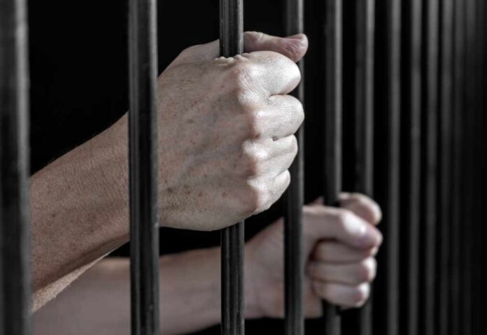 Prisoner's,Hands,Gripping,The,Bars,Of,A,Cell,In,Jail