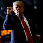 A man in a suit with a red tie raises his fist in a confident gesture against a dark background