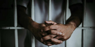 A close-up of a persons hands clasped together in front of prison bars
