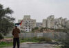 Massive Displacement: A West Bank Emergency Person holding a Palestinian flag in front of buildings and a barrier