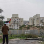 Person holding a Palestinian flag in front of buildings and a barrier