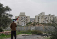 Person holding a Palestinian flag in front of buildings and a barrier