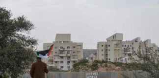 Person holding a Palestinian flag in front of buildings and a barrier