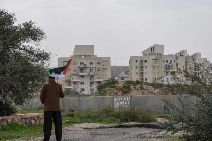 Person holding a Palestinian flag in front of buildings and a barrier