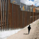 A person walking along a border wall in a desert area under a cloudy sky