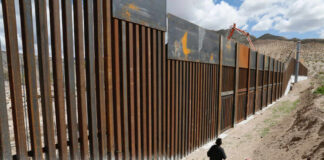 A person walking along a border wall in a desert area under a cloudy sky