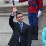 A man in a suit waving during a public event, accompanied by a woman in a blue outfit