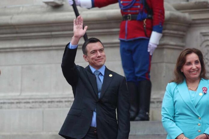 A man in a suit waving during a public event, accompanied by a woman in a blue outfit
