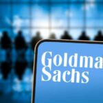 A smartphone displaying the Goldman Sachs logo in front of a blurred group of people in a meeting