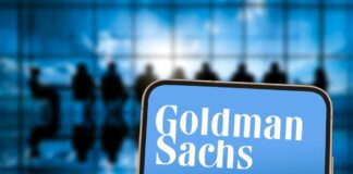 A smartphone displaying the Goldman Sachs logo in front of a blurred group of people in a meeting