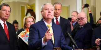 A group of government officials at a press conference with one speaking into a microphone