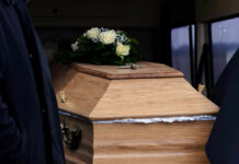A wooden coffin adorned with white roses and greenery, accompanied by a person in dark clothing