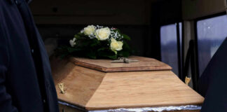 A wooden coffin adorned with white roses and greenery, accompanied by a person in dark clothing