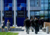 Military personnel walking past NATO banners with two women conversing in the foreground
