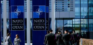 Military personnel walking past NATO banners with two women conversing in the foreground