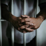 A close-up of a persons hands clasped together in front of prison bars