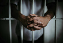 A close-up of a persons hands clasped together in front of prison bars
