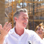 A man speaking at a construction site with a microphone, smiling, while a woman in the background laughs.