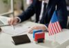 An official reviewing documents with a small American flag and stamps on the table
