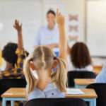 Children in a classroom raising their hands to answer a question while a teacher stands in front