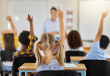 Children in a classroom raising their hands to answer a question while a teacher stands in front