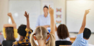 Children in a classroom raising their hands to answer a question while a teacher stands in front