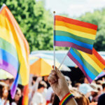 A hand holding rainbow flags at a pride event with a crowd in the background