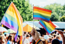 A hand holding rainbow flags at a pride event with a crowd in the background