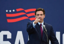 A man in a suit pointing while speaking into a microphone against a backdrop featuring an American flag design