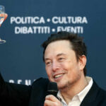 A man in a black suit raising a glass of water while smiling at an event