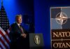 A man speaking at a podium with a NATO logo and an American flag in the background