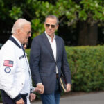 Two men walking outdoors, one in a jacket with an American flag, the other in a blazer
