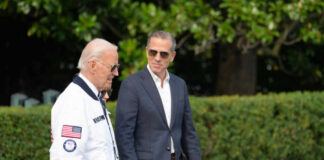 Two men walking outdoors, one in a jacket with an American flag, the other in a blazer