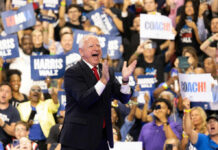 A man in a suit enthusiastically claps at a political rally with a cheering crowd holding signs