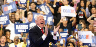 A man in a suit enthusiastically claps at a political rally with a cheering crowd holding signs