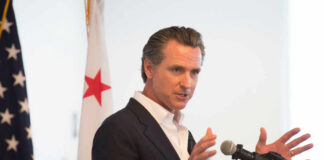 A man in a suit speaking into a microphone with a California flag in the background