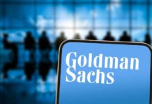 Goldman Rides War Wave—Stunning Profit Spike A smartphone displaying the Goldman Sachs logo in front of a blurred group of people in a meeting
