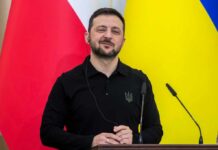 A political leader speaking at a press conference with flags in the background