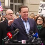 A politician speaking at a rally with supporters and protest signs in the background