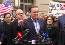 A politician speaking at a rally with supporters and protest signs in the background