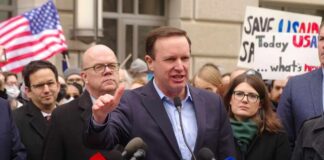 A politician speaking at a rally with supporters and protest signs in the background