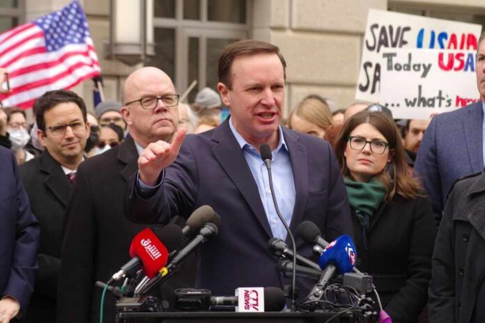 A politician speaking at a rally with supporters and protest signs in the background