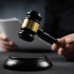 A gavel held above a sounding block with a person reading documents in the background