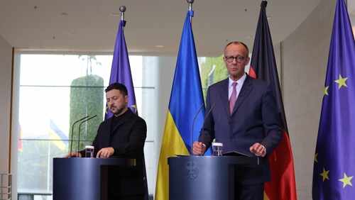 shutterstock_2633921363.jpg Two government officials at a press conference with flags in the background