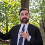 Man in a suit speaking outdoors with trees in the background