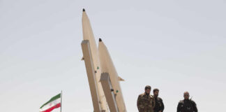 Military personnel standing near missile launchers with an Iranian flag in the background