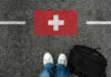 Immigration LIMIT: Switzerland’s 10 Million Dilemma Feet wearing white sneakers standing on a road with a painted Swiss flag and a black backpack nearby