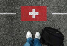 Feet wearing white sneakers standing on a road with a painted Swiss flag and a black backpack nearby