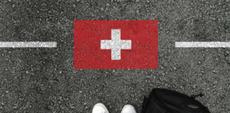 Feet wearing white sneakers standing on a road with a painted Swiss flag and a black backpack nearby