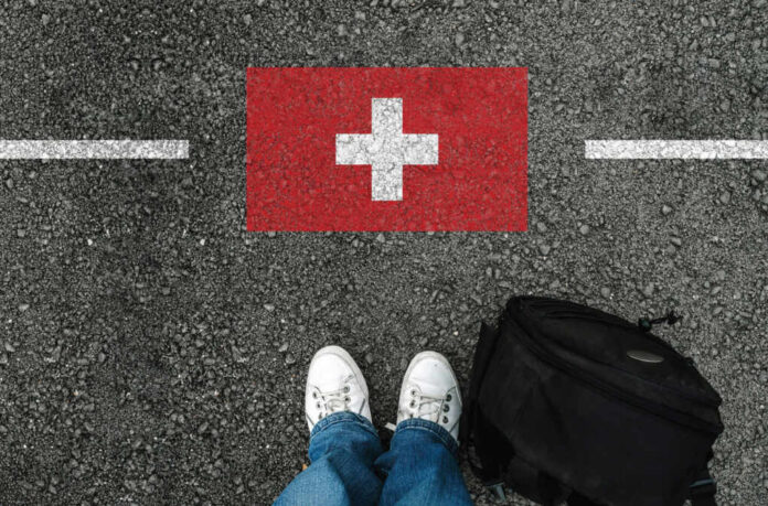 Feet wearing white sneakers standing on a road with a painted Swiss flag and a black backpack nearby